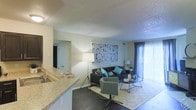 This single-floor space showcases an open-concept design that seamlessly connects the kitchen and living area, creating a cohesive and functional layout. The kitchen features granite-style countertops and a sink positioned at a breakfast bar that overlooks the living room, making it ideal for both everyday use and entertaining.

The living area is generously proportioned, accommodating a full seating arrangement with a sofa, accent chair, and coffee table. A textured ceiling adds architectural character throughout the space, while dark hardwood-style flooring grounds the interior with a warm, contemporary feel. A large abstract wall piece serves as a focal point above the seating area.

Natural light is a notable feature, with floor-to-ceiling vertical blinds flanking a sliding glass door that opens to an outdoor area, brightening the living space considerably. A floor lamp provides additional ambient lighting for the room.

The layout also includes a hallway leading to additional rooms, suggesting a practical flow throughout the unit. The overall ambiance is modern and welcoming, with neutral wall tones complemented by vibrant accent furnishings in teal and green hues.
