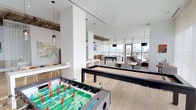 This contemporary amenity space is thoughtfully designed with an open, flowing layout that seamlessly connects multiple distinct zones. Light hardwood flooring runs throughout, unifying the space while complementing the predominantly white palette of smooth and brick-textured wall surfaces.

The game area is a central highlight, featuring a foosball table and a sleek black billiards table positioned beneath modern pendant lighting. A dedicated lounge zone offers clusters of stylish grey upholstered armchairs arranged around low coffee tables, ideal for relaxed conversation. A separate seating area includes a comfortable sofa grouping, partially framed by a striking mixed-material room divider combining reclaimed wood and dark metal framing, adding warmth and visual texture.

A casual dining and café-style section provides round and rectangular tables with upholstered seating. A bar-height counter with wooden stools offers additional casual seating near what appears to be a kitchenette or service area.

Floor-to-ceiling windows on multiple sides flood the space with abundant natural light and provide sweeping elevated city views. Sheer drapery softens the light while maintaining the airy atmosphere. Direct access to an outdoor terrace further enhances the open, connected feel of this well-appointed communal space.