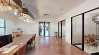 Located at 1582 Dogwood Dr in Oxford, Ohio, this thoughtfully designed single-floor workspace combines rustic charm with contemporary finishes to create a functional and visually engaging environment. The space is unified by richly stained polished concrete floors that add warmth and character throughout, complemented by crisp white shiplap walls that lend a clean, modern-farmhouse aesthetic.

The main open area features a substantial reclaimed wood conference table paired with leather chairs, anchored beneath a striking statement chandelier crafted from a repurposed galvanized barrel fitted with Edison-style bulbs — a standout focal point that sets the tone for the space's eclectic design sensibility. A dedicated workstation area is equipped with desktop computers atop a solid wood desk, also bathed in the warm glow of pendant Edison bulbs overhead.

Glass-partitioned rooms with sleek black frames provide enclosed meeting or breakout spaces without sacrificing visual openness. One such room features a round dining-style table with vibrant orange chairs and a layered chandelier, set atop patterned tile flooring. Natural light filters in through multiple windows, while access to adjacent areas is provided through a combination of dark wood doors and black-framed glass doors. The overall ambiance is professional yet inviting, balancing industrial, rustic, and modern influences seamlessly.
