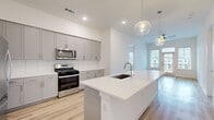 This well-designed 2-bedroom, 2-bathroom single-floor home offers a clean, modern aesthetic throughout. The open-concept kitchen serves as the heart of the space, featuring gray shaker-style cabinetry, a subway tile backsplash, stainless steel appliances including a gas range and built-in microwave, and a large center island with a white quartz countertop, undermount sink, and built-in shelving. Globe pendant lights hang above the island, adding a contemporary touch. Light hardwood-style flooring flows through the main living areas, complemented by recessed lighting and ceiling fans. The living area benefits from natural light and access to an outdoor balcony. Both bedrooms are carpeted and include ceiling fans, with the primary bedroom featuring an en-suite bathroom. The primary bath is finished with large-format tile flooring, gray vanity cabinetry, a white quartz countertop, and a marble-surround soaking tub. Each bedroom is supported by a dedicated closet, with the primary walk-in closet offering wrap-around shelving and hanging rods. A hallway connects the rooms efficiently, and two louvered closet doors near the kitchen provide additional storage. The overall palette of white walls, soft neutrals, and brushed gold and silver fixtures creates a bright, cohesive living environment.