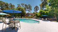 Located in Columbia, South Carolina, this outdoor pool area offers a well-appointed recreational retreat surrounded by mature trees and manicured landscaping. The centerpiece is a generously sized freeform swimming pool with crystal-clear blue water, accessible via stainless steel entry rails and accented by decorative lane markers along the pool floor. The expansive concrete sun deck encircles the pool, providing ample space for lounging and socializing. Numerous reclining lounge chairs with teal cushions are positioned throughout the deck, offering guests comfortable spots to relax in the South Carolina sun. Multiple blue market umbrellas and canopy structures are strategically placed to provide shaded seating options, complemented by high-top bistro tables and chairs for casual outdoor dining. A pool house structure anchors one side of the deck, featuring large windows and direct access to the patio area, with additional covered seating nearby. The surrounding landscape is rich with tall pine trees and leafy deciduous trees, creating a natural privacy screen and a serene, park-like atmosphere. Neatly trimmed shrubs and ornamental plantings line the perimeter fencing, adding color and texture to the environment. The two-story residential buildings visible in the background provide context for the community setting, completing this inviting outdoor amenity space.