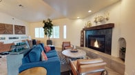 This single-floor space offers a thoughtfully designed living area that blends rustic charm with modern comfort. The centerpiece of the room is a bold fireplace framed by a substantial wood mantel and a sleek surround, above which decorative sculptural pieces — including animal mounts and artful accents — add a distinctive, nature-inspired character. Vaulted ceilings lend an open, airy quality to the space, while recessed lighting provides a clean, unobtrusive source of illumination throughout. The living area is furnished with plush teal sofas accented by coral throw pillows, complemented by rich cognac leather chairs that reinforce the warm, eclectic aesthetic. A textured round stone coffee table anchors the seating arrangement atop a geometric patterned area rug. A tall indoor tree adds a natural, organic element near the bright, shuttered windows, which allow for controlled natural light. The warm, neutral wall tones throughout create a cohesive backdrop that ties the varied textures and materials together. Overall, this space strikes a welcoming balance between rustic décor and contemporary design sensibilities, making it a functional and visually engaging environment.