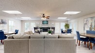 Located at 2601 W 12th St in Russellville, Arkansas, this single-floor space offers a well-appointed, open layout designed for comfort and functionality. The main living area is a standout, featuring light wood-style flooring, a textured ceiling with multiple skylights and a ceiling fan, and a classic fireplace framed by a white mantle. Large windows along the front wall flood the space with natural light and offer pleasant outdoor views of mature trees, contributing to a warm and inviting ambiance. A wall-mounted television above the fireplace anchors the room, complemented by ample seating and tasteful decorative artwork. The dining area is situated nearby and is accessible through elegant French doors, adding a refined architectural touch. Multiple dining configurations are present throughout, making the space well-suited for group use. The kitchen features tile flooring, white cabinetry, dark countertops, and a peninsula with bar seating, providing a practical yet stylish workspace. Throughout the space, neutral tones and contemporary décor create a cohesive, modern aesthetic. The combination of natural light, thoughtful layout, and versatile areas makes this a comfortable and highly functional space.