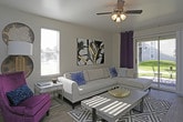 This single-floor space showcases a well-appointed living area designed with both comfort and style in mind. The living room serves as the focal point, featuring neutral-toned walls that provide a versatile backdrop for the bold, contemporary furnishings. A large L-shaped sectional sofa in light gray anchors the space, complemented by striking navy and teal accent pillows that add a vibrant pop of color. A plush purple accent chair introduces an eclectic touch, reinforcing the room's confident use of color.

Natural light filters in through multiple windows fitted with white blinds, brightening the space and highlighting the smooth, light-toned flooring. A ceiling fan with an integrated light fixture is centrally positioned overhead, providing both functionality and a finishing architectural detail. Deep navy curtain panels frame a glass door, adding drama while connecting the interior to an outdoor area.

Wall décor includes a bold abstract canvas print and a pair of decorative circular wall pieces, contributing to the room's curated, modern aesthetic. The overall ambiance is contemporary and inviting, balancing neutral foundations with rich jewel-toned accents throughout.