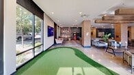 Located at 9310 Otis Ave in Lawrence, Indiana, this single-floor community amenity space offers a generously sized, open-concept layout designed for recreation, relaxation, and socializing. Large floor-to-ceiling windows line multiple walls, flooding the interior with abundant natural light and providing views of the surrounding outdoor areas.

The space is thoughtfully divided into distinct zones. An indoor putting green with artificial turf serves as a standout recreational feature, stretching across a polished concrete floor near the main entrance. Adjacent to this is a game lounge area outfitted with a shuffleboard table, a classic arcade machine, and comfortable seating arrangements set against patterned carpeting and a dark accent wall.

A well-appointed kitchen area showcases teal cabinetry, stainless steel appliances, a central island with bar seating, and decorative pendant lighting suspended from exposed wood beam accents on the ceiling. Unique rope-hung sculptural figures adorn the walls, adding an artistic touch throughout the space.

The lounge area features a large sectional sofa, ottoman, and a dining table with seating, complemented by wall-mounted televisions and decorative wall art. Exposed brick columns punctuate the open floor plan, adding warmth and architectural character. A secured glass door leads to an on-site fitness center, rounding out this well-equipped community hub.