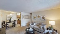 This single-floor residence offers a welcoming and functional layout designed for comfortable everyday living. The living room serves as the heart of the home, featuring plush wall-to-wall carpeting in a warm neutral tone that adds softness underfoot. A textured popcorn ceiling adds a classic touch overhead, while the walls are painted in a soft beige hue that creates a bright and inviting atmosphere. Decorative wall art in golden floral tones and geometric wood-framed panels add personality and warmth to the space. The living area comfortably accommodates a full seating arrangement, centered around a dark round coffee table that anchors the room. The space flows naturally into a dedicated dining area, where a dark dining set is positioned adjacent to the kitchen. The kitchen features medium-toned wood cabinetry and is equipped with standard appliances, offering a practical and efficient workspace. A partial wall separates the kitchen from the main living area, maintaining an open feel while providing subtle definition between spaces. The overall ambiance throughout is cozy, cohesive, and move-in ready, with warm finishes and thoughtful décor creating a harmonious and livable environment.