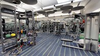 This fitness facility offers a comprehensive workout environment designed to accommodate a variety of training styles and fitness goals. The space is generously sized, providing ample room between equipment stations to allow users to move freely and safely during their workouts.

The strength training area is extensively outfitted with a diverse selection of selectorized weight machines, including cable systems and multi-functional units such as the Synergy360, which supports suspension and functional training exercises. Free weights, including kettlebells and medicine balls, are neatly organized along one side of the floor, catering to those who prefer dynamic, free-movement training.

The facility features bright, even overhead lighting supplemented by distinctive circular acoustic panels suspended from the ceiling, which help manage sound levels throughout the space. Full-length mirrors line the walls, enhancing the sense of openness while allowing users to monitor their form during exercise.

The flooring consists of a durable blue carpet with linear detailing, providing both comfort underfoot and a clean, professional aesthetic. The overall ambiance is functional and well-organized, reflecting a facility built to support serious, consistent training.