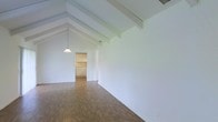 This single-story space makes a strong architectural impression with its vaulted ceiling and exposed white-painted beams that follow the roofline, creating a sense of height and openness rarely found in similarly scaled properties. The dramatic pitch of the ceiling draws the eye upward and contributes to an expansive, airy feel throughout the main living area. A pendant light hangs centrally from the ceiling, adding a simple, functional accent that complements the clean, minimalist aesthetic of the space. The walls are finished in crisp white, enhancing the brightness of the interior, while the warm-toned laminate or vinyl plank flooring grounds the space and adds a natural contrast to the light surroundings. An open passageway connects the main living area to the kitchen, which features white cabinetry and appears well-integrated into the overall floor plan. The layout flows naturally between spaces, making the interior feel cohesive and practical. The overall ambiance is clean, open, and inviting, with the vaulted beamed ceiling serving as the defining architectural feature of the space.
