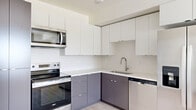This efficiently designed single-floor unit offers a thoughtfully laid-out interior comprising a kitchen, living room, and bathroom. The kitchen is a standout feature, showcasing a two-tone cabinet scheme with sleek white upper cabinets and charcoal-gray lower cabinets, complemented by white countertops and a full suite of stainless steel appliances, including a range, over-the-range microwave, dishwasher, and a side-by-side refrigerator. A single-basin undermount sink with a modern arc faucet anchors the workspace. Light wood-look flooring runs seamlessly throughout the kitchen and into the open living area, creating a cohesive and airy feel. The living room is bright and open, with clean white walls, neutral tones, and a window fitted with horizontal blinds that allows natural light to fill the space. The bathroom features a dark-cabinetry vanity with a white countertop and undermount sink, brushed nickel light fixtures above, and wood-look flooring that ties the space together. Clean lines, neutral finishes, and contemporary hardware define the overall aesthetic, resulting in a modern and functional living environment.