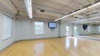 This single-floor studio facility is designed to accommodate a variety of active and multipurpose uses. The main studio area is expansive and open, featuring polished light hardwood flooring that extends throughout, creating a clean, bright aesthetic underfoot. The soaring ceilings are left exposed, showcasing industrial-style steel trusses, ductwork, and overhead fluorescent strip lighting that floods the space with consistent, even illumination.

The studio walls are finished in a neutral light gray, providing a calm, functional backdrop. A full wall of mirrors along one side enhances the sense of depth and is particularly well-suited for fitness or dance instruction. Wall-mounted flat-screen displays, paired with a projection surface, support instructional or presentation needs. Yoga mats stored along the perimeter indicate the room's adaptability for wellness programming.

Natural light enters through multiple windows, offering views of the surrounding exterior. Glass-panel doors connect the studio to an adjacent workspace or administrative area, visible through the transparent partitions and equipped with desks and office seating. The overall ambiance is modern, functional, and highly adaptable, making this space an excellent fit for group fitness classes, dance rehearsals, wellness sessions, or community programming.