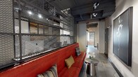 Business Center 2 is a single-floor commercial space that masterfully blends industrial design with contemporary style. The space is defined by its exposed ceiling structure, featuring dark-painted beams and overhead metal grid shelving units that lend a raw, urban character throughout. Track lighting is strategically mounted above, casting focused illumination across the space and highlighting its key features.

A standout element is the bold red leather sofa anchoring the lounge area, accompanied by an industrial-style metal side table adorned with a floral accent. The seating area is positioned alongside metal mesh partition shelving, which serves both as functional storage and a distinctive architectural divider.

The walls display striking large-format black-and-white photographic artwork featuring smoke imagery, adding an artistic and editorial edge to the environment. A corridor connects to additional areas of the space, where large multi-pane windows allow natural light to filter through, offering a contrast to the otherwise moody interior palette.

Polished concrete-style flooring runs throughout, complementing the industrial aesthetic, while textured area rugs add warmth underfoot. Exposed brick visible in the background further reinforces the space's character. Overall, Business Center 2 delivers a professional yet creatively charged environment suited for collaborative or client-facing business use.