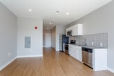 This single-floor unit at 1600 Genessee St in Kansas City, Missouri offers a clean, modern design with an open-concept layout that integrates the living and kitchen areas seamlessly. Warm-toned hardwood flooring runs throughout, complementing the neutral gray and white walls to create a bright, cohesive atmosphere.

The kitchen is well-appointed with stainless steel appliances, including a range, over-the-range microwave, dishwasher, and refrigerator. Glossy white cabinetry pairs with sleek countertops and a classic white subway tile backsplash, adding a contemporary aesthetic. Recessed lighting throughout the main living area enhances the overall brightness of the space.

Large sliding glass doors and adjacent windows allow generous natural light to fill the living area while providing access to an outdoor space and views beyond. The open floor plan offers flexibility for various furniture arrangements.

The bathroom features wood-look flooring that carries through from the main living areas, and includes a spacious walk-in shower enclosed by a full glass sliding door with chrome fixtures, contributing to the clean, modern feel of the unit. The overall design reflects a practical yet stylish approach suited for everyday living.