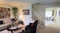 Model 6441 is a two-story residence that offers a well-organized and comfortable layout spread across two levels. The main living area greets visitors with warm, neutral-toned walls and plush carpeting underfoot, creating an inviting atmosphere suited for relaxing or entertaining. The space is furnished with stylish seating arrangements, including accent chairs with nail-head detailing, and a round glass-top coffee table that adds a touch of elegance to the room. A framed piece of wall art serves as a focal point, complemented by soft ambient lighting from a table lamp and a wall-mounted sconce. A tall potted plant in the corner introduces a natural element, enhancing the overall warmth of the space. The carpeted staircase, outfitted with a white metal railing, connects the two floors with ease and serves as a central architectural feature of the home. A glimpse beyond the staircase reveals a dining area bathed in natural light from nearby windows, suggesting a bright and airy quality throughout. The two-floor configuration provides a clear separation between living and private spaces, making this model an efficient and practical home with thoughtful design details throughout.