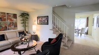 Model 6441 is a two-story residence that offers a well-organized and comfortable layout spread across two levels. The main living area greets visitors with warm, neutral-toned walls and plush carpeting underfoot, creating an inviting atmosphere suited for relaxing or entertaining. The space is furnished with stylish seating arrangements, including accent chairs with nail-head detailing, and a round glass-top coffee table that adds a touch of elegance to the room. A framed piece of wall art serves as a focal point, complemented by soft ambient lighting from a table lamp and a wall-mounted sconce. A tall potted plant in the corner introduces a natural element, enhancing the overall warmth of the space. The carpeted staircase, outfitted with a white metal railing, connects the two floors with ease and serves as a central architectural feature of the home. A glimpse beyond the staircase reveals a dining area bathed in natural light from nearby windows, suggesting a bright and airy quality throughout. The two-floor configuration provides a clear separation between living and private spaces, making this model an efficient and practical home with thoughtful design details throughout.
