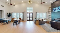Located at 4739 Starr Ranch Rd in Indian Land, SC, this single-floor community clubhouse is designed to impress with its dramatic vaulted ceilings and an abundance of natural light streaming through large windows on all sides. The main lounge area serves as the heart of the space, anchored by a sleek floor-to-ceiling tile fireplace wall with built-in shelving and a mounted television, complemented by a curated arrangement of comfortable sofas and accent chairs atop a woven area rug. Light hardwood flooring runs throughout, creating a warm, cohesive aesthetic. At the entrance, soaring cathedral ceilings and double glass doors welcome visitors, with an elegant chandelier and ceiling fans adding both style and function overhead. A casual dining nook features a tufted caramel leather banquette, marble-topped bistro tables, and an eye-catching wall display of sculptural ceramic art pieces, while a wall-mounted game board adds a playful, social touch. The kitchen bar area is outfitted with a generous quartz-topped island with seating, dark cabinetry, open shelving, a striking geometric tile accent wall, and a mounted TV — ideal for casual gatherings. Together, these zones create a versatile, modern communal retreat with a sophisticated yet inviting atmosphere.
