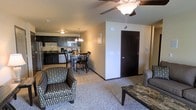 This single-floor space offers a thoughtfully designed open-concept layout that seamlessly connects the living, dining, and kitchen areas into one cohesive living environment. The living area features plush carpeting, a ceiling fan with integrated lighting, and comfortable seating arranged around a dark-toned coffee table, creating a relaxed and inviting atmosphere. Transitioning naturally from the living space, the dining area accommodates a small table with chairs, lit overhead by a classic chandelier that adds a touch of warmth to the space. The kitchen is well-equipped with dark cabinetry, a full suite of appliances including a microwave, stove, and dishwasher, and a double sink set into a dark countertop. Light tile flooring in the kitchen and dining area provides a clean contrast to the carpeted living zone. A dark-stained interior door leads to additional areas of the unit, maintaining a consistent design aesthetic throughout. The warm beige walls and layered lighting — from the chandelier to the ceiling fan fixture — contribute to a cozy, functional ambiance that makes this space both practical and comfortable for everyday living.