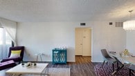 This single-floor residence presents a straightforward and functional layout designed for comfortable everyday living. The living area serves as the heart of the space, featuring dark hardwood-style flooring that adds warmth and contrast against the crisp, neutral white walls. A textured popcorn ceiling runs throughout, lending a classic finish to the interior. The room receives ample ambient light, contributing to an open and airy feel. A central HVAC vent integrated into the ceiling ensures year-round climate comfort. The living space is well-proportioned, accommodating a seating arrangement alongside accent furniture, including a distinctive teal cabinet with lattice-patterned glass doors that adds a pop of color and personality to the room. A simple, clean coffee table and patterned area rugs define the living zone, while the layout flows naturally toward an interior doorway, hinting at additional rooms beyond. The transition between spaces is marked by a clean archway and a white interior door, maintaining a cohesive and uncluttered aesthetic throughout. Overall, this space combines practicality with understated charm, making it a welcoming and adaptable environment.