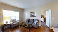 This single-floor space is designed with a practical and comfortable layout. The living room serves as the central gathering area, featuring warm hardwood flooring that adds character and a sense of warmth throughout. Neutral, beige-toned walls create a calm and inviting atmosphere, complemented by white trim and clean architectural lines. A window along one wall allows natural light to filter into the room, brightening the space during the day. The living area comfortably accommodates a full sofa and accent chair arrangement, centered around a classic wooden coffee table, making it well-suited for relaxed everyday living. A framed piece of wall art adds a decorative focal point above the sofa. A doorway visible from the living room provides a direct connection to a carpeted bedroom, which features a wooden dresser and a neatly made bed, suggesting a private and restful retreat. The transition between the hardwood living area and the carpeted bedroom highlights a thoughtful use of materials to distinguish functional zones. Overall, the space conveys a tidy, functional, and welcoming environment suitable for comfortable day-to-day living.