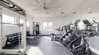 Located at 43808 Vintage St in Lancaster, CA, this dedicated exercise room offers a comprehensive fitness facility designed for a full range of workouts. The space features crisp white walls, durable rubber flooring, and recessed lighting complemented by a large ceiling fan that ensures comfort during workouts. Natural light streams in through multiple windows on two walls, providing views of the surrounding neighborhood and creating an open, airy atmosphere. Full-length mirrors line one wall, enhancing the sense of space and allowing users to monitor their form. The cardio section is equipped with treadmills, StairMaster steppers, a stationary bike, and a spin bike, while the strength training area includes a multi-function cable machine, a power rack, a flat/adjustable weight bench, a dumbbell rack with a full set of free weights, and additional plate-loaded equipment. A water fountain is conveniently mounted near the exit door, adding a practical amenity for users. The clean, minimalist design keeps the focus on functionality, making this exercise room a well-rounded fitness facility suitable for both cardio and strength training.
