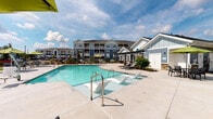 Located at 3963 Woolcock Dr in Myrtle Beach, South Carolina, this single-story amenity space serves as a central gathering hub within a well-appointed residential community. The exterior architecture combines classic coastal design with refined material choices, most notably the striking stacked stone accent walls that incorporate rich tones of rust, tan, and charcoal. These stone elements are paired with blue-gray horizontal lap siding and crisp white trim, creating a visually cohesive and polished aesthetic.

The centerpiece of the outdoor environment is a generously sized swimming pool with clear, turquoise water, accessible via sleek entry steps and handrails. The expansive concrete pool deck is outfitted with modern chaise lounges and wicker seating with neutral cushions, providing ample space for relaxation under the open South Carolina sky.

Adjacent to the pool, a covered patio offers a shaded lounge area furnished with comfortable wicker seating arrangements, ideal for socializing or unwinding. Stacked stone columns anchor the covered structure, reinforcing the architectural character seen throughout the space. White-paned glass doors with grid detailing provide entry points and allow natural light to filter through. Lush landscaping and manicured greenery frame the perimeter, enhancing the overall resort-like ambiance of this outdoor amenity space.