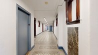 This single-floor corridor showcases a thoughtful blend of classic architectural detailing and functional design. The hallway stretches generously, with a floor-plan model revealing an intersecting layout that connects multiple wings, providing efficient circulation throughout the building.

The most striking feature underfoot is the ornate mosaic tile flooring, composed of small penny tiles arranged in elaborate geometric and floral patterns. The intricate designs run the length of the corridor, serving as a decorative centerpiece that speaks to the building's historic character.

Lining both walls, dark mahogany-stained door surrounds and transom frames provide rich contrast against the crisp white walls and blue baseboard trim. The unit doors themselves are painted in a muted steel blue, complementing the overall color palette. Decorative wall niches framed in matching dark wood millwork punctuate the corridor at regular intervals, adding architectural depth.

Crown molding runs along the ceiling junctions in portions of the hallway, reinforcing the classical sensibility of the space. Overhead, linear fluorescent lighting fixtures provide consistent, even illumination throughout. A marble-clad feature element is visible near the elevator lobby area, adding another layer of refined material detail to this well-preserved and characterful shared corridor.