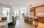 This single-floor residence showcases a thoughtfully designed open-concept layout that seamlessly connects the kitchen, dining, and living areas. The kitchen is outfitted with two-tone cabinetry combining walnut wood and white upper cabinets, a stainless steel refrigerator, built-in appliances, and a stone-topped island with seating for casual dining. An angular pendant light fixture adds a striking design element overhead. Light wood flooring flows throughout, contributing to a clean, cohesive aesthetic.

The living area benefits from floor-to-ceiling windows that frame urban views and flood the space with natural light. The bedroom maintains the same crisp, white palette, featuring a window with city views, wall-mounted sconce lighting, and a sliding door for privacy.

Two bathrooms serve the unit — one featuring a glass-enclosed shower with marble-style tile walls and dark metal framing, accompanied by a wood-panel vanity with granite countertop. The second bathroom similarly features a glass shower enclosure with refined finishes. Throughout the space, the consistent use of white walls, light flooring, and dark metal accents creates a polished, contemporary ambiance.