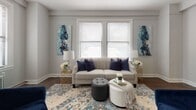 This single-floor residence combines classic pre-war character with updated finishes across a well-proportioned layout. The living room serves as the heart of the home, bathed in natural light from multiple double-hung windows fitted with blinds. Rich, dark hardwood floors run throughout, providing a warm contrast to the neutral gray walls and crisp white trim. Cast-iron radiators underscore the building's architectural heritage. The living area offers generous space for seating arrangements and includes a dedicated media wall with convenient access to an adjoining area.

The bedroom is comfortably scaled, featuring the same hardwood flooring and a window with a radiator beneath it, ensuring both natural light and warmth. A second, more compact bedroom or flex room provides additional versatility.

The bathroom has been thoughtfully updated, showcasing floor-to-ceiling marble-style porcelain tile with dramatic veining in white and gray tones. A soaking tub with a tiled surround, white cabinetry, and a small window add brightness and elegance to the space.

Overall, the residence offers a cohesive blend of pre-war charm and modern refinement, with abundant natural light and classic details throughout.