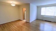 This single-floor residence offers a clean, functional layout with a warm and inviting atmosphere. The living and dining areas flow seamlessly together, unified by smooth hardwood flooring in a light honey tone that runs throughout the space, adding warmth and a sense of continuity. Crisp white walls provide a bright, neutral backdrop that enhances the natural light filtering in through the windows.

A central partial wall divides the main living area from the adjacent dining space, creating a sense of separation while maintaining an open feel. The dining area is accented by a ceiling-mounted light fixture, providing ambient overhead lighting.

The kitchen is accessible through a doorway and is distinctly defined from the main living space. It features warm wood cabinetry in an upper configuration and is equipped with a full-size refrigerator, offering practical storage and functionality. The kitchen's compact layout makes efficient use of its footprint.

Throughout the unit, clean baseboards frame the hardwood floors, and multiple electrical outlets are conveniently placed along the walls. The window in the secondary area allows natural light to brighten the space, contributing to the overall light and airy ambiance of this well-maintained, straightforward residential unit.