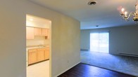 This single-floor space offers a practical and comfortable layout with distinct living and kitchen areas. The kitchen, visible through an open archway, is outfitted with light wood cabinetry providing ample storage, a countertop with a built-in sink, and neutral-toned finishes that give the space a clean, understated look. The smooth white ceiling and neutral wall tones carry throughout, creating a cohesive aesthetic.

The living area extends from the kitchen zone and is carpeted, contrasting with the dark hardwood-style flooring in the entryway and transition space. A large sliding glass door with vertical blinds anchors the far wall of the living area, allowing abundant natural light to fill the room and offering a view of the outdoors. A baseboard heater sits beneath the window, providing functional heating along the exterior wall.

The ceiling features a centrally placed ventilation unit, and the overall layout flows naturally from the kitchen through to the living space. The neutral palette and open feel make this a versatile space suitable for a variety of furnishing arrangements.