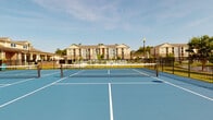 Located at 6710 Wahoo Dr in Myrtle Beach, South Carolina, this outdoor recreational facility features multiple well-maintained pickleball courts surfaced in a vivid blue hard court finish with crisp white line markings and regulation nets. The courts are fully enclosed by sturdy black wrought iron fencing, providing both safety and an open, airy feel that allows for unobstructed views of the surrounding landscape.

The setting is notably scenic, with lush green lawns, mature trees, and a tranquil pond visible just beyond the court perimeter. The backdrop includes an attractive multi-story residential community featuring traditional architectural detailing, brick accents, and covered balconies. A lamppost stands nearby, indicating the courts are accessible during evening hours as well.

The courts benefit from abundant natural light under open South Carolina skies, making for an inviting and energetic atmosphere for players of all skill levels. The overall environment is clean, well-kept, and thoughtfully integrated into the broader community grounds, offering a seamless connection between active recreation and the natural surroundings. This amenity reflects a strong commitment to an active outdoor lifestyle within a well-appointed residential setting.