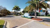 Bridgewater is a single-story residential community that makes a striking first impression with its beautifully landscaped entrance. The community is framed by an abundance of tall, swaying palm trees and verdant tropical greenery that evokes a distinctly Florida lifestyle. A welcoming arched monument sign serves as the gateway to the neighborhood, set against manicured mulched garden beds and well-maintained grounds. The entrance features decorative paver brickwork that guides residents and visitors along a clean, organized roadway, reflecting attention to detail in the community's design. The architecture visible from the entry is characteristic of Florida residential construction, with light-toned exteriors that complement the bright, sunny surroundings. The overall ambiance of Bridgewater is one of a peaceful, well-kept community that balances tropical charm with neighborhood accessibility. The lush landscaping and mature trees create natural shade and privacy throughout the grounds, contributing to a serene and inviting atmosphere that is emblematic of South Florida living.