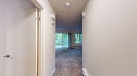 Located at 930 Sunrise Pl SW in Issaquah, Washington, this single-level unit is nestled within a verdant, tree-lined setting that defines much of its character. The layout flows from an entry hallway with a textured ceiling through to the main living area, where plush carpeting runs underfoot and natural light pours in through a large sliding glass door and an adjacent picture window. Both openings frame sweeping views of dense Pacific Northwest woodland, creating a serene, nature-immersed atmosphere throughout the living space. The sliding door opens directly onto a private balcony enclosed by a white railing, offering an unobstructed view of lush greenery and mature trees. The kitchen is efficiently laid out with white cabinetry extending to the ceiling, light-toned countertops, a built-in cooktop, a sink, and a black refrigerator, all set over warm hardwood-style flooring. The bedroom features soft carpeting, a mirrored sliding-door closet with built-in shelving, and access to an adjoining room. The bathroom is clean and bright, finished with wood-style flooring, a white vanity, and a wall-mounted heater. Textured ceilings and baseboard heating are consistent throughout the unit.