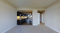 This single-floor space features an open-concept design that seamlessly connects the kitchen and living area. The kitchen is outfitted with dark espresso cabinetry that contrasts sharply against light-toned walls, creating a bold, contemporary aesthetic. Stainless steel appliances, including a gas range, built-in microwave, and refrigerator, are integrated throughout. The kitchen also features a granite-style countertop, an undermount sink with modern fixtures, and recessed under-cabinet lighting that adds warmth to the workspace. A pendant light fixture hangs above, serving as a subtle focal point. The flooring transitions from tile in the kitchen to plush carpet in the living area, clearly delineating the two zones while maintaining a cohesive feel. The living space is generously proportioned, with smooth walls and neutral tones that create a calm, versatile backdrop. A hallway visible in the background suggests additional rooms beyond, with recessed ceiling lighting providing consistent illumination. Overall, the space offers a clean, functional, and modern environment suited to contemporary living.