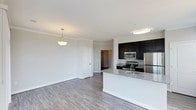 This single-floor residence offers a thoughtfully designed open-concept layout that seamlessly connects the kitchen and living areas, creating a bright and functional living environment. The kitchen is a standout feature, showcasing rich dark cabinetry that contrasts elegantly with light granite countertops. A central island with a built-in sink provides additional prep space and serves as a natural gathering point. Stainless steel appliances, including a gas range, over-the-range microwave, and refrigerator, complete the kitchen's modern and practical appeal. Flush ceiling lighting keeps the kitchen well-illuminated, while a decorative pendant light adds warmth to the adjacent living area. The living space is open and airy, with neutral-toned walls and light hardwood-style flooring that flows consistently throughout, lending the home a cohesive and contemporary feel. Crown molding adds an elegant architectural detail along the ceiling line. A white interior door leads to additional spaces within the home, hinting at further functional areas beyond the main living zone. The overall ambiance is clean, modern, and move-in ready, with a balanced mix of stylish design and everyday practicality.