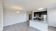 This single-floor residence offers a thoughtfully designed open-concept layout that seamlessly connects the kitchen and living areas, creating a bright and functional living environment. The kitchen is a standout feature, showcasing rich dark cabinetry that contrasts elegantly with light granite countertops. A central island with a built-in sink provides additional prep space and serves as a natural gathering point. Stainless steel appliances, including a gas range, over-the-range microwave, and refrigerator, complete the kitchen's modern and practical appeal. Flush ceiling lighting keeps the kitchen well-illuminated, while a decorative pendant light adds warmth to the adjacent living area. The living space is open and airy, with neutral-toned walls and light hardwood-style flooring that flows consistently throughout, lending the home a cohesive and contemporary feel. Crown molding adds an elegant architectural detail along the ceiling line. A white interior door leads to additional spaces within the home, hinting at further functional areas beyond the main living zone. The overall ambiance is clean, modern, and move-in ready, with a balanced mix of stylish design and everyday practicality.