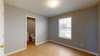 This single-floor space is finished in a consistent, neutral gray palette throughout, creating a calm and cohesive aesthetic across all rooms. The flooring is uniform warm-toned ceramic tile, laid in a grid pattern that extends seamlessly from room to room, contributing to a clean and practical interior.

The main room is generously proportioned, featuring gray walls trimmed with crisp white baseboards and a textured ceiling with a central flush-mount light fixture. A window fitted with horizontal blinds allows natural light to filter into the space, brightening the room during daylight hours. Multiple electrical outlets are positioned along the walls, offering functional convenience.

An adjoining room of similar size and finish includes two interior doors — one leading to a closet and another opening directly into the bathroom. This layout provides a convenient and connected flow between living and private spaces.

The bathroom is straightforwardly appointed, featuring a white vanity with a single sink and mirror, a standard toilet, and a full-size tub and shower combination enclosed by white surrounds. The tile flooring continues into this space, maintaining design consistency.

Overall, the space presents a clean, functional layout with a neutral, move-in ready finish suitable for a variety of uses.