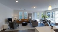 This single-floor space showcases a thoughtfully designed open-concept layout that seamlessly blends living and dining areas into one cohesive, inviting environment. The living area is anchored by a rich tan leather sofa paired with contemporary gray armchairs and a tufted ottoman coffee table, all arranged atop a soft area rug over dark hardwood flooring that runs throughout the space. Recessed lighting complements the natural light streaming in through multiple double-hung windows, giving the interior a bright and airy feel. A set of floor-to-ceiling sliding glass doors opens directly to an outdoor patio area, framed by lush greenery that brings a refreshing natural element indoors. The dining area, positioned adjacent to the glass doors, features a mid-century modern table and chairs, making it ideal for both casual meals and entertaining. The neutral color palette of soft whites and grays creates a clean, modern ambiance, while carefully selected accent lighting and furnishings add warmth and character. The overall atmosphere is sophisticated yet comfortable, with an open flow and generous sense of space that makes the layout feel both functional and welcoming.