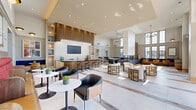 Located at 600 West State Street in Kennett Square, PA, this single-floor community lounge offers a generously proportioned, open-plan gathering space designed with contemporary sophistication. The layout is organized into several distinct functional zones that flow seamlessly into one another, creating a dynamic yet cohesive environment.

At the heart of the space is a sleek wet bar featuring a dark stone countertop, wood-toned cabinetry with vertical slat detailing, a mounted flat-screen display, and bar-height seating — ideal for social gatherings. Adjacent to the bar, a central island with a striking blue countertop provides additional prep and serving space.

The lounge area is anchored by generous modular seating in warm amber and navy tones, complemented by round bistro tables and modern accent chairs. A games corner houses a full-sized pool table set beneath soaring double-height ceilings and floor-to-ceiling multi-pane windows that flood the space with abundant natural light and offer open views of the surrounding Kennett Square streetscape.

Warm wood flooring, a vertical wood slat partition, recessed lighting, and a sleek fireplace contribute to the refined yet approachable ambiance. The large-format tile near the entry transitions gracefully to wood flooring throughout the lounge, reinforcing the thoughtful material palette that defines this well-appointed communal space.