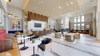 Located at 600 West State Street in Kennett Square, PA, this single-floor community lounge offers a generously proportioned, open-plan gathering space designed with contemporary sophistication. The layout is organized into several distinct functional zones that flow seamlessly into one another, creating a dynamic yet cohesive environment.

At the heart of the space is a sleek wet bar featuring a dark stone countertop, wood-toned cabinetry with vertical slat detailing, a mounted flat-screen display, and bar-height seating — ideal for social gatherings. Adjacent to the bar, a central island with a striking blue countertop provides additional prep and serving space.

The lounge area is anchored by generous modular seating in warm amber and navy tones, complemented by round bistro tables and modern accent chairs. A games corner houses a full-sized pool table set beneath soaring double-height ceilings and floor-to-ceiling multi-pane windows that flood the space with abundant natural light and offer open views of the surrounding Kennett Square streetscape.

Warm wood flooring, a vertical wood slat partition, recessed lighting, and a sleek fireplace contribute to the refined yet approachable ambiance. The large-format tile near the entry transitions gracefully to wood flooring throughout the lounge, reinforcing the thoughtful material palette that defines this well-appointed communal space.