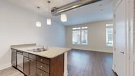 Located at 5350 Burnet Rd in Austin, Texas, this single-floor unit offers a thoughtfully designed open-concept layout that maximizes every square foot. The combined kitchen and living area serves as the heart of the space, anchored by a peninsula-style kitchen island topped with granite countertops and fitted with rich dark wood cabinetry and a double stainless steel sink. A trio of modern cube pendant lights hang overhead, while an exposed industrial HVAC duct adds a loft-inspired character to the ceiling. The living area flows naturally from the kitchen and features durable hardwood-style flooring throughout, neutral light gray walls, and a large window that invites ample natural light. A separate room, ideal as a bedroom or flex space, includes a ceiling fan with light, tall windows with blinds for adjustable light control, and the same warm hardwood-style flooring. The full bathroom features tile flooring, a granite-topped vanity with dark wood cabinetry, a tub-shower combination, and clean white walls that create a bright, functional space. A dedicated closet provides convenient built-in storage. This unit combines modern finishes with an efficient, well-organized floor plan in a prime Austin location.