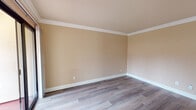 This single-floor space is thoughtfully laid out to maximize functionality across its living areas. Throughout the main living spaces, grey-toned hardwood-style flooring provides a clean, cohesive aesthetic, complemented by warm beige walls and crisp white crown molding and baseboards that add a polished architectural detail to every room.

The living area is a generous, open space anchored by a fireplace, creating a natural focal point. A sliding glass door opens directly onto an outdoor patio, allowing natural light to fill the room while offering views of the surrounding exterior. The bedroom features plush carpeting and a full-width mirrored closet with built-in shelving and a hanging rod, providing ample storage. Crown molding continues into this room, maintaining a consistent design language throughout.

The kitchen is designed for efficiency, featuring granite-style countertops, grey cabinetry on both sides, a stainless steel sink, and white appliances including a refrigerator and electric range. Light hardwood-style flooring carries through the kitchen as well. The bathroom is finished with large-format tile walls and flooring, and includes a walk-in shower with frosted sliding glass doors and brushed nickel hardware, offering a clean and functional design.