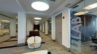 This single-floor office space presents a polished, contemporary design that balances functionality with aesthetic appeal. The main corridor serves as a central artery, lined with a combination of private offices enclosed by full-height glass partitions and solid doors, offering both visual connectivity and acoustic privacy. Large, circular flush-mounted ceiling lights are evenly distributed along the hallway, casting a clean, even illumination throughout the space. The flooring consists of patterned commercial carpet in neutral tones, contributing to a professional yet welcoming atmosphere. A built-in wooden reception or workstation counter runs along one side of the corridor, providing a refined focal point finished in warm walnut-toned cabinetry. Seating areas are integrated naturally into the flow of the corridor, featuring modern, curved white lounge chairs paired with compact side tables, ideal for informal meetings or brief waiting areas. The walls are crisp white, enhancing the sense of openness, while branded glass panels add a bold graphic element that reinforces a corporate identity. Overall, the space exudes a well-organized, professional ambiance suited for a dynamic office environment.