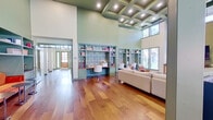 This thoughtfully designed single-floor space makes a striking impression through its dramatic, soaring ceiling heights and cohesive sage-green color palette carried throughout every room. The main living area centers on a statement fireplace console with patterned cabinet doors, a wall-mounted television, and flanking sconce lighting. Seating includes a pair of cognac leather lounge chairs with ottomans and a neutral sofa, all anchored on area rugs atop richly toned wide-plank hardwood floors. Clerestory windows flood the space with natural light while offering views of the surrounding outdoors.

A dedicated study zone features an expansive wall of sage-green built-in shelving accented by botanical-patterned wallpaper, with dual built-in wooden desks and paired swivel chairs providing functional workspace. Nearby, a vibrant burnt-orange banquette with open shelving above adds a bold accent and additional seating. The kitchenette is outfitted with sage-green shaker cabinetry, dark stone countertops, a herringbone-tile backsplash, a farmhouse sink, wine refrigerator, and brass pendant lighting. A large picture window within the kitchen offers direct views to an outdoor patio area, enhancing the connection between interior and exterior.