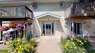 This single-floor unit is situated within a multi-story residential building that presents a tidy, well-maintained exterior. The building features light-colored lap siding and a distinctive peaked entryway canopy marked with the address number 307, creating a clear and inviting focal point for arrivals. A concrete walkway leads directly to the glass-paneled entry doors, lined on both sides by neatly kept garden beds bursting with yellow day lilies and other seasonal plantings that add color and curb appeal to the approach.

Flanking the central entrance are upper-level balconies with wood railings, indicating shared building amenities above. The exterior also features wall-mounted air conditioning units, suggesting climate-controlled comfort within each unit. The building's overall design is practical and residential in character, with a clean, understated aesthetic that emphasizes functionality.

As the only available visual documentation pertains to the building's exterior, interior details of the single-floor unit are not available for description. However, the building's well-kept grounds and thoughtfully landscaped entry suggest a consistently maintained living environment throughout.