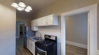 This single-floor space showcases a well-appointed kitchen as its central feature. The kitchen is equipped with stainless steel appliances, including a gas range with black accents, a refrigerator, and a double-basin sink with a modern faucet. White upper and lower cabinetry provides ample storage, complemented by neutral-toned walls that create a clean, cohesive aesthetic. A white tile backsplash adds a classic touch along the lower portion of the walls, while hardwood-style flooring runs throughout, adding warmth and continuity to the space. An elegant multi-bulb pendant light fixture overhead provides functional and decorative illumination. The kitchen connects seamlessly to an adjacent room visible through a doorway, suggesting an open and accessible floor plan. An arched architectural detail is visible along one wall, lending a subtle decorative character to the overall design. The space features crisp white trim and molding throughout, contributing to a tidy, well-finished appearance. Overall, the layout and finishes create a practical yet inviting environment suitable for everyday living.