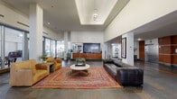 This lobby in New Orleans, Louisiana, presents a carefully designed entry experience that balances functionality with contemporary aesthetics. The space is organized into distinct zones, beginning with a corridor-style entry wing characterized by large floor-to-ceiling glass panels that allow abundant natural light to filter in, offering views of the exterior. Multicolored slate tile flooring runs throughout, lending a grounded, textural quality to the overall design. Warm wood-panel wall accents complement the neutral white walls and ceilings, creating a cohesive visual identity.

The central gathering area features soaring ceiling heights, anchored by a raised coffered section with pendant lighting. This zone is furnished with leather seating, a colorful braided area rug, and a round coffee table, providing a welcoming lounge environment. A front desk finished in warm wood tones serves as the operational hub, and decorative artwork is displayed throughout, adding character to the space.

The elevator bank is a notable architectural feature, with multiple stainless steel elevator doors framed by rich wood surrounds. Adjacent transitional corridors connect the various functional areas, including service counters and restroom access, ensuring a logical and efficient flow through the lobby.