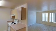 This single-floor residence offers a straightforward and functional layout designed for comfortable everyday living. The kitchen is equipped with a white electric range, a built-in overhead exhaust hood, and light wood cabinetry providing ample storage space. Countertops offer practical workspace, and the kitchen opens directly toward the living area, creating a connected and accessible flow between the two spaces. The living area is bright and open, benefiting from natural light that enters through a window fitted with vertical blinds. The space features clean white walls and light-toned ceramic tile flooring, which extends consistently throughout the unit, lending a cohesive and easy-to-maintain finish. A hallway leads from the main living area to the remaining rooms, lined with wall-mounted light fixtures that provide warm illumination. HVAC vents are integrated into the ceilings, ensuring climate control throughout. The overall design is clean and neutral, offering a blank canvas that is well-suited for a variety of furnishing styles. The unit's simple, efficient layout makes it a practical and livable space from entry to end.