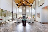 Located in Layton, Utah, this remarkable single-floor living space makes an immediate impression with its dramatic, vaulted cathedral ceiling supported by exposed natural wood trusses — a striking architectural centerpiece visible from every angle of the room. The soaring ceiling height amplifies the sense of openness and grandeur throughout the space.

The living room is bathed in natural light, courtesy of a generous arrangement of clerestory windows positioned high along the walls, as well as large lower windows that frame views of the surrounding outdoor greenery. The floor is finished in large-format stone-look tiles that complement the space's clean, modern aesthetic.

A floor-to-ceiling stacked stone fireplace with a bold, rustic wood beam mantel serves as a commanding focal point, adding warmth and texture to the otherwise contemporary interior. Adjacent to the main seating area, a frameless glass partition wall with an etched mountain landscape design elegantly defines a secondary lounge area while preserving the open, airy flow of the space.

The walls feature curated large-format nature photography, nodding to Utah's scenic landscapes and reinforcing the connection to the outdoors. A decorative oversized wall clock adds an artistic touch near the entryway. Together, these elements create a sophisticated, welcoming atmosphere that is both architecturally distinctive and functionally refined.