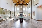 Located in Layton, Utah, this remarkable single-floor living space makes an immediate impression with its dramatic, vaulted cathedral ceiling supported by exposed natural wood trusses — a striking architectural centerpiece visible from every angle of the room. The soaring ceiling height amplifies the sense of openness and grandeur throughout the space.

The living room is bathed in natural light, courtesy of a generous arrangement of clerestory windows positioned high along the walls, as well as large lower windows that frame views of the surrounding outdoor greenery. The floor is finished in large-format stone-look tiles that complement the space's clean, modern aesthetic.

A floor-to-ceiling stacked stone fireplace with a bold, rustic wood beam mantel serves as a commanding focal point, adding warmth and texture to the otherwise contemporary interior. Adjacent to the main seating area, a frameless glass partition wall with an etched mountain landscape design elegantly defines a secondary lounge area while preserving the open, airy flow of the space.

The walls feature curated large-format nature photography, nodding to Utah's scenic landscapes and reinforcing the connection to the outdoors. A decorative oversized wall clock adds an artistic touch near the entryway. Together, these elements create a sophisticated, welcoming atmosphere that is both architecturally distinctive and functionally refined.