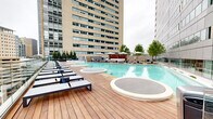 This sophisticated rooftop amenity space delivers a resort-caliber experience within an urban setting, offering a seamless blend of relaxation and city living. The centerpiece is a generously sized outdoor swimming pool with crisp turquoise water, flanked by warm hardwood decking that extends across the terrace. Rows of modern chaise lounges are arranged along the deck, providing ample space for sunbathing while taking in panoramic views of the surrounding downtown skyline through glass perimeter railings that preserve unobstructed sightlines. Shade umbrellas and additional seating areas are positioned poolside, offering both sun and shelter options. Curated planters filled with manicured trees and greenery provide natural privacy screens and soften the urban environment. A dedicated indoor lounge area opens directly onto the terrace, featuring contemporary furnishings including woven lounge chairs, a long upholstered banquette positioned beneath expansive windows, and accent lighting embedded in the ceiling that creates a warm, polished ambiance. Bold staircase access connects the terrace levels, finished with red treads and stainless railings against a tile plaza surround, reinforcing the space's clean, modern aesthetic throughout.